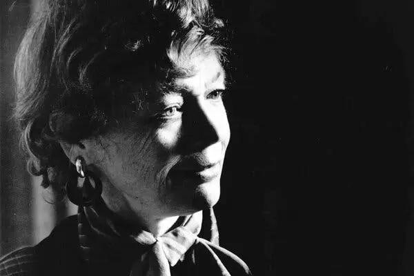 یک عکس سیاه‑سفید متین از صورت زن به صورت نزدیک. موهای کوتاه و موجدار دارد، گوشوارهی حلقهای و یک شال ابریشمی در گلویش گره خورده است. به سمت راست قاب نگاه میکند و نیمهخندیده است.