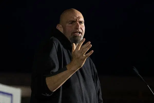 عکسی از مردی کچل در یک سوئیتشرتم تاریک، دست راست بالا، دهان باز بهصورت بیانگر، در پسزمینهٔ تاریک.