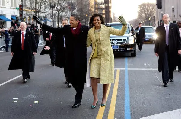 باراک و میشل اوباما در حالی که خوشحال می‌خندند و دست در دست قدم می‌زنند، در ایندِرسن‌اِوِنیو. او یک لباس و کت زرد کم‌رنگ به تن دارد؛ او کت تیره و شال رنگ شرابی پوشیده است. مأموران سرویس مخفی در کنارشان قدم می‌زنند.
