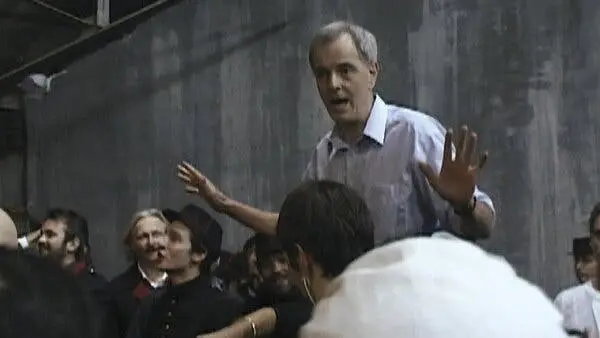 مردی با پیراهن آبی روشن، دست‌هایش را بالا برده و در صحنه‌ی فیلم‌برداری به گروهی از افراد لباس‌دار زمان قدیم که برخی خندان به او نگاه می‌کنند، صحبت می‌کند.