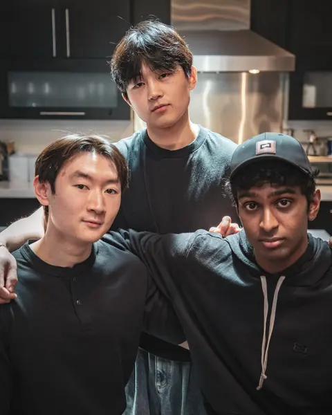 سه مرد جوان که پیراهن‌های تاریک پوشیده‌اند، در یک آشپزخانه در کنار هم ایستاده‌اند. یکی ایستاده و دو نفر دیگر نشسته‌اند، در حالی که بازوهایشان به‌هم گره خورده است، زیر نور نرم.