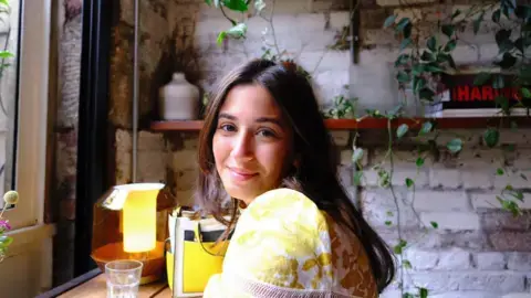 دختری با موهای قهوه‌ای و پیراهن زرد در یک کافی‌شاپ نیمه‌تاریک لبخند می‌زند.