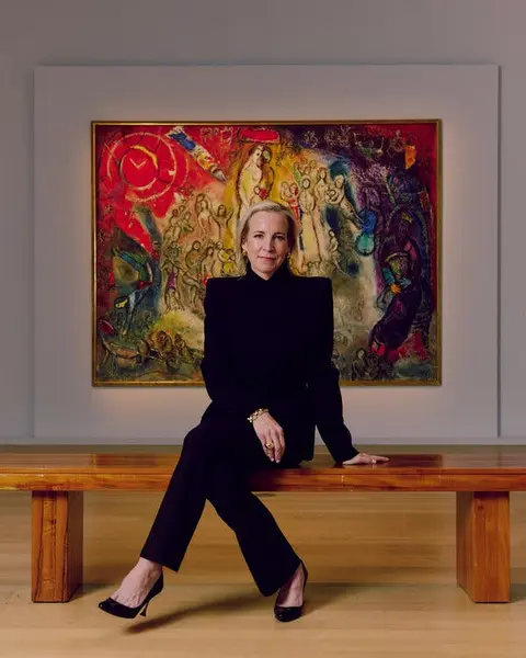 بونی برنان، که لباس مشکی پوشیده است، بر روی نیمکتی چوبی صیقلی در مقابل یک نقاشی بسیار رنگارنگ در گالری با دیوارهای سفید نشسته است.