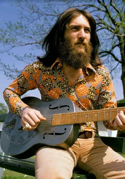 استیو کروپر جوان، با موهای بلند و تیره و ریش قهوه‌ای، پیراهن چاپی و شلوار روشن، گیتاری در دست دارد و به دوردست نگاه می‌کند.