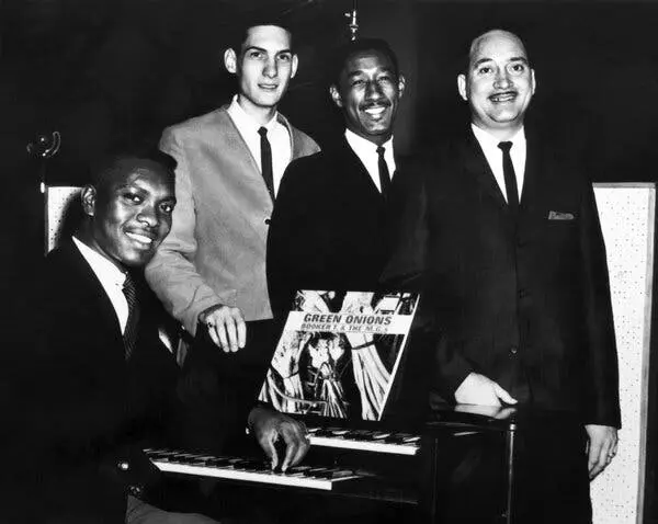 عکس سیاه‌سفید چهار مرد جوان، کت‌وشلوار پوشیده و کراوات‌های باریک و تیره، دور یک کیبورد جمع شده‌اند.