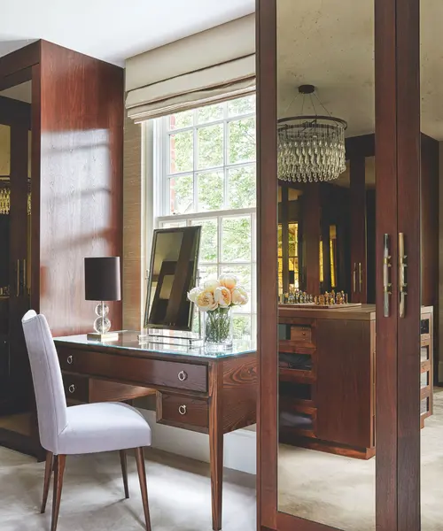کمدهای قهوهای آینهای در اتاقخواب با یک میز آرایش قهوهای آینهای، صندلی سفید، آینه و پنجرهٔ بزرگ روشن با پردههای بژ